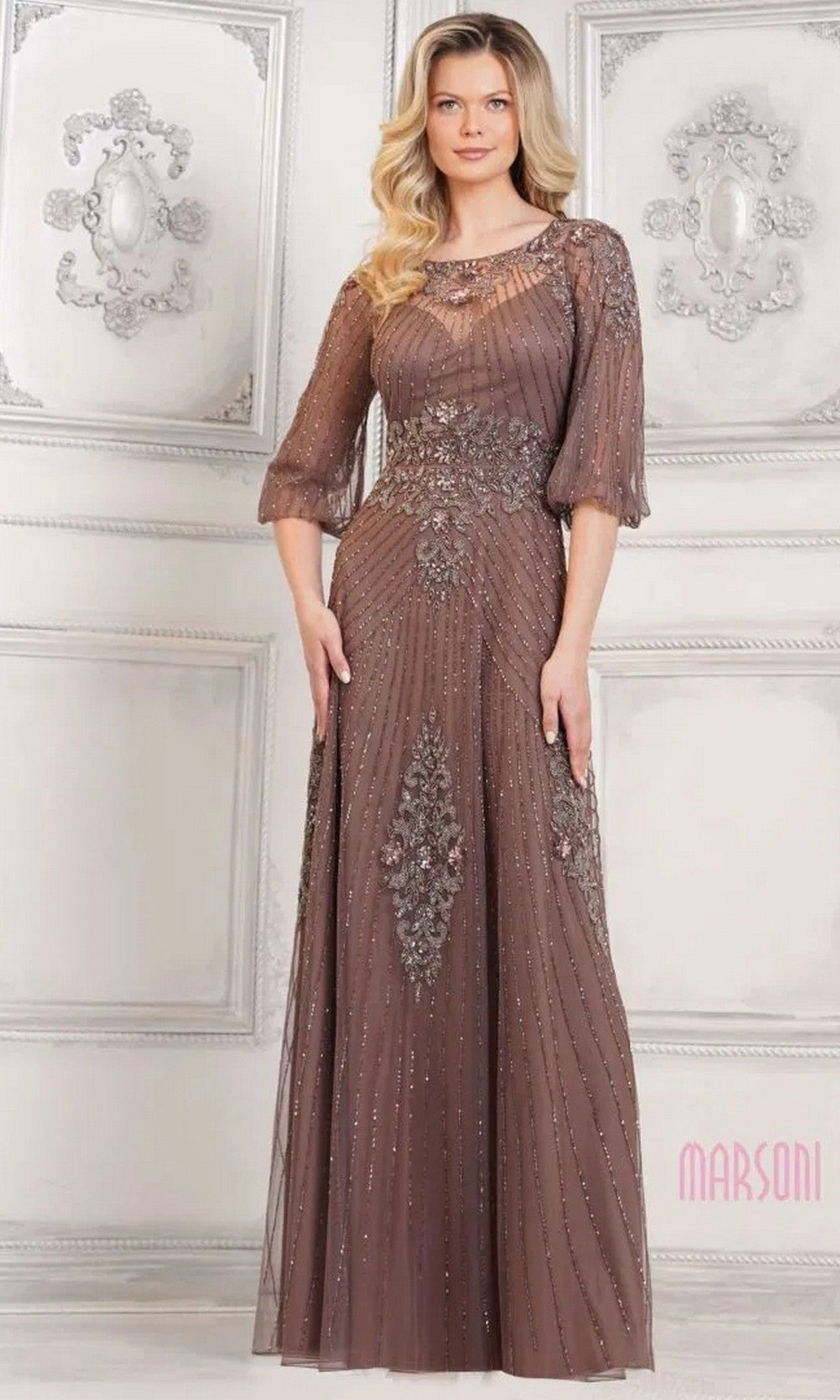 A woman in a floor-length, dark taupe Long Formal Dress: Marsoni MV1323 with sheer sleeves and intricate beadwork stands against an ornate, light-colored wall.